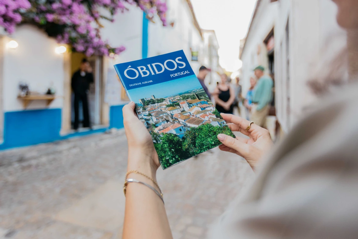 Visitante segura um guia turístico de Óbidos enquanto explora as ruas históricas da vila medieval de Óbidos, em Portugal, rodeada por casas tradicionais e ambiente turístico.