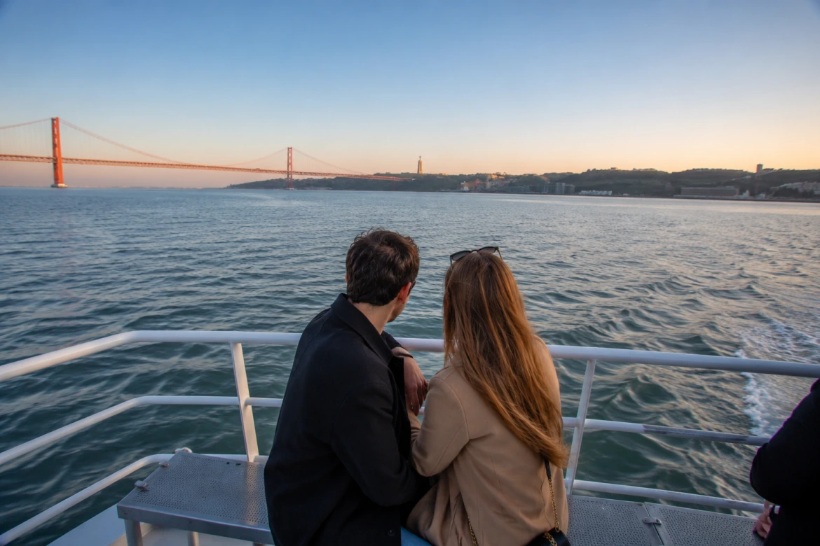 Casal a desfrutar de um cruzeiro ao pôr do sol no Rio Tejo com vista para a Ponte 25 de Abril e o Cristo Rei em Lisboa