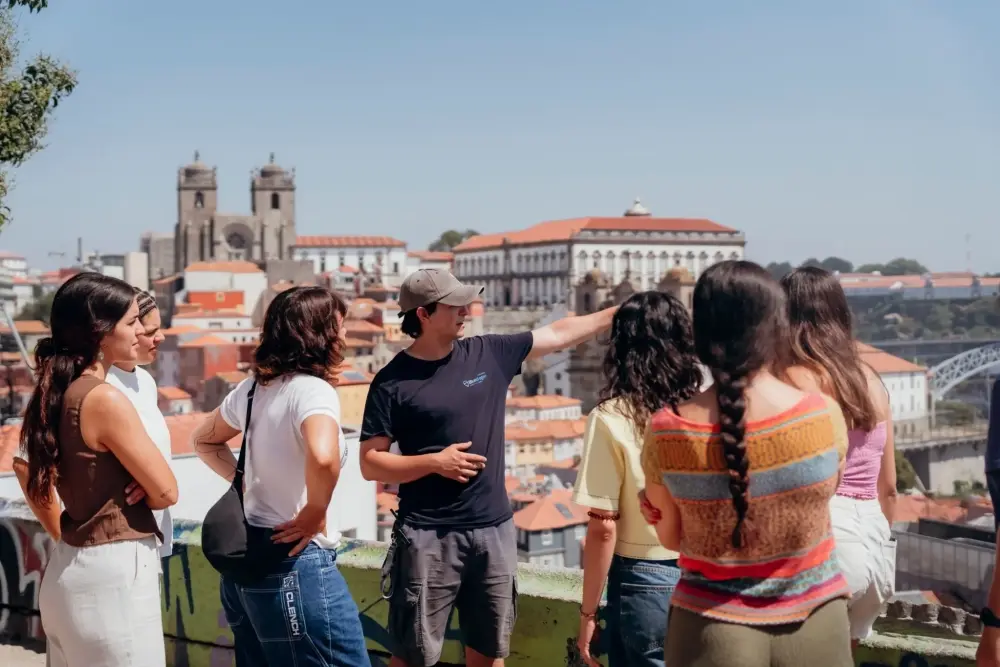 Guia fala com visitantes num miradouro do Porto, com a Sé do Porto, os telhados do centro histórico e a Ponte D. Luís I ao fundo.