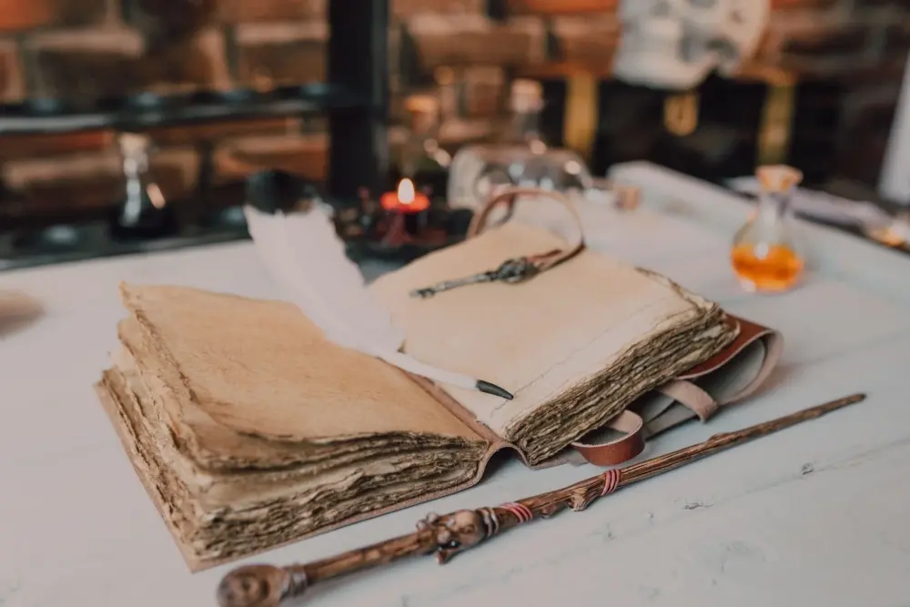 Livro de estilo antigo aberto com uma pena branca, chave decorativa, varinha e pequeno frasco de vidro sobre uma mesa.