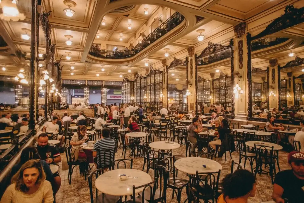 Interior ornamentado do Café Majestic, no Porto, com paredes espelhadas, tetos decorativos e mesas ocupadas por visitantes.