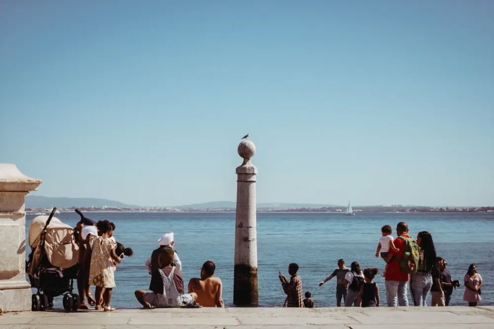 People relaxing by the Tagus River at Cais das Colunas in Lisbon, with a stone column and sailboat in view.