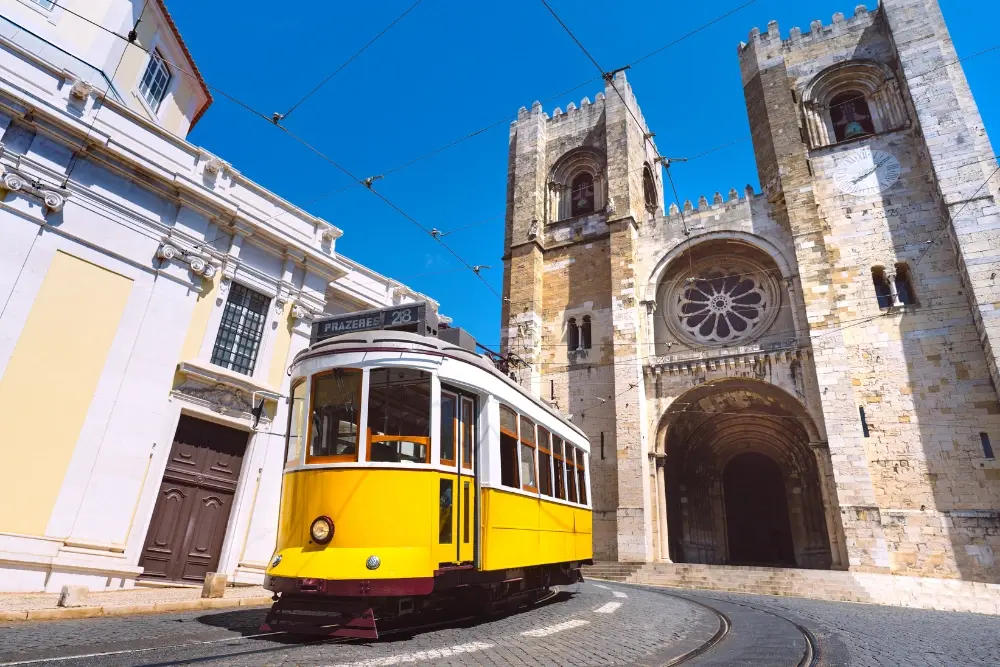 Elétrico 28 amarelo passa em frente à Sé de Lisboa, com as torres e a rosácea da catedral ao fundo.