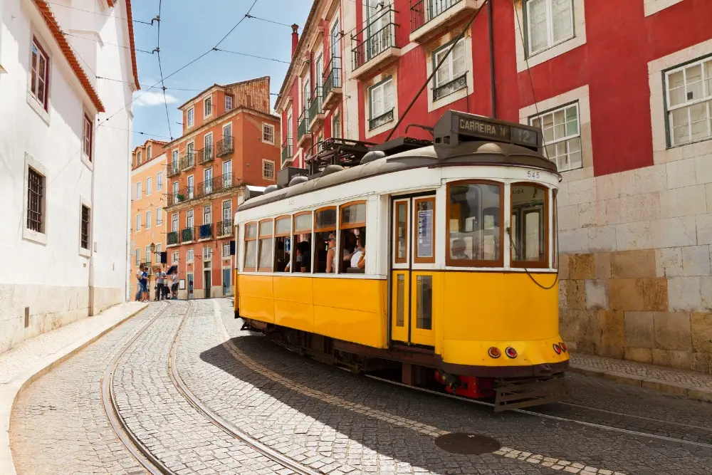 Elétrico tradicional amarelo percorre uma rua estreita e empedrada entre edifícios coloridos em Alfama, Lisboa.